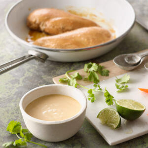 Ginger Soy Marinade In a ramekin. It is made was Mountain High Plain Yoghurt, pineapple juice, soy sauce, and ginger root. On the left of the ramekin, there is a frying pan with three chicken marinated chicken breasts. Next to the pan, there is a cutting board with some measuring spoons and garnishments such as cilantro, lime, and sliced carrot. Beside the cutting board, there is a knife.