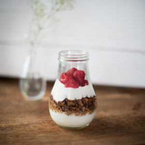 A Yoghurt Parfait in a glass jar is shown. It contains a layer of Mountain High Vanilla Yoghurt, a layer of granola, then another layer of Mountain High Vanilla Yoghurt, and a final top layer of fresh berries.